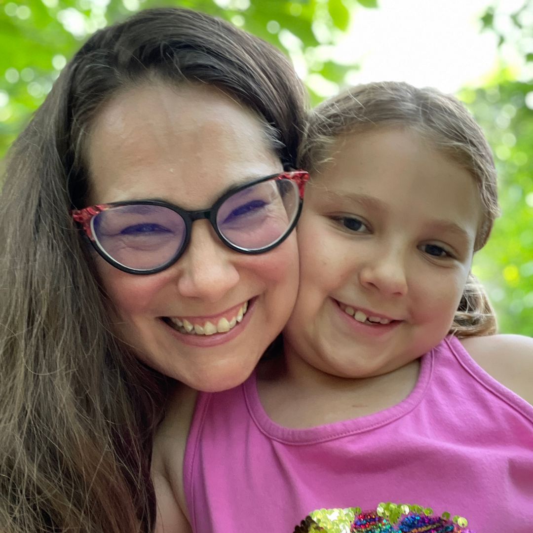 A photo of a middle-aged white woman and a young white child. The woman is smiling at the camera. She wears black and red glasses and has long brown hair. The young kid is also smiling at the camera and wearing a pink shirt. Their hair is pulled back from their face. There are green leaves in the background. 
